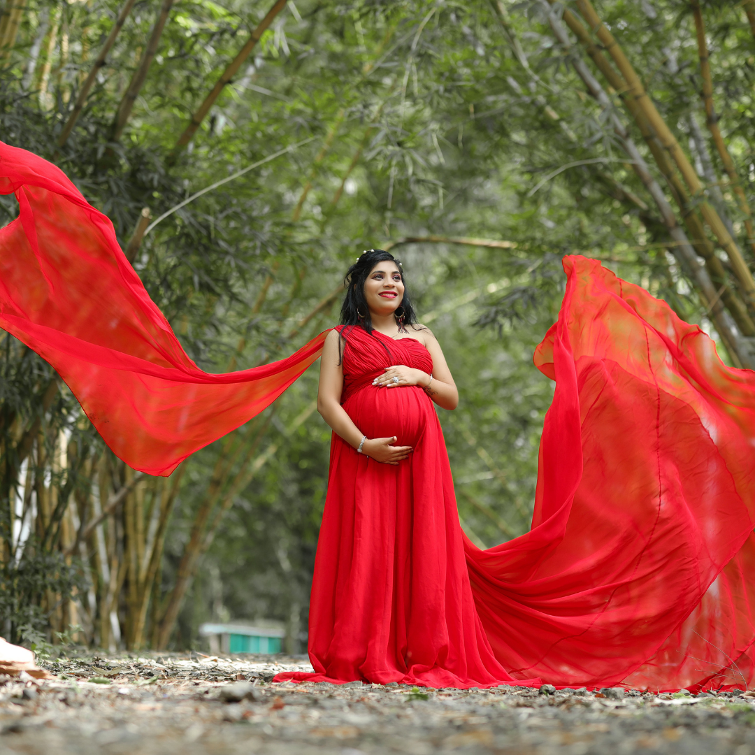 Pregnant woman holding her belly. Standing between trees on a cloudy day covered by soft light. She stands wearing a beautiful flowing red maternity gown. Her hair and makeup are done to match the outfit and she is glowing with joy!