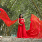 Pregnant woman holding her belly. Standing between trees on a cloudy day covered by soft light. She stands wearing a beautiful flowing red maternity gown. Her hair and makeup are done to match the outfit and she is glowing with joy!