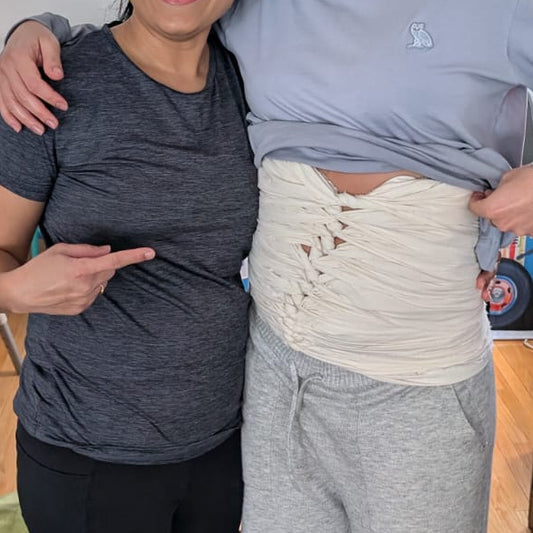 Two women posing together in a living room. The woman on the right is wearing a light blue long-sleeve shirt, gray sweatpants, and a beige maternity support belt worn over her stomach.